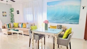 Un salon avec une table basse à plateau de verre, un canapé agrémenté de coussins colorés et un grand tableau représentant une vague océanique accroché au mur.