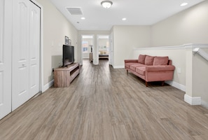 Un vaste espace de vie doté d’un plancher en bois, d’un canapé rose, d’un téléviseur posé sur un meuble en bois, et d’un couloir menant à une autre pièce.