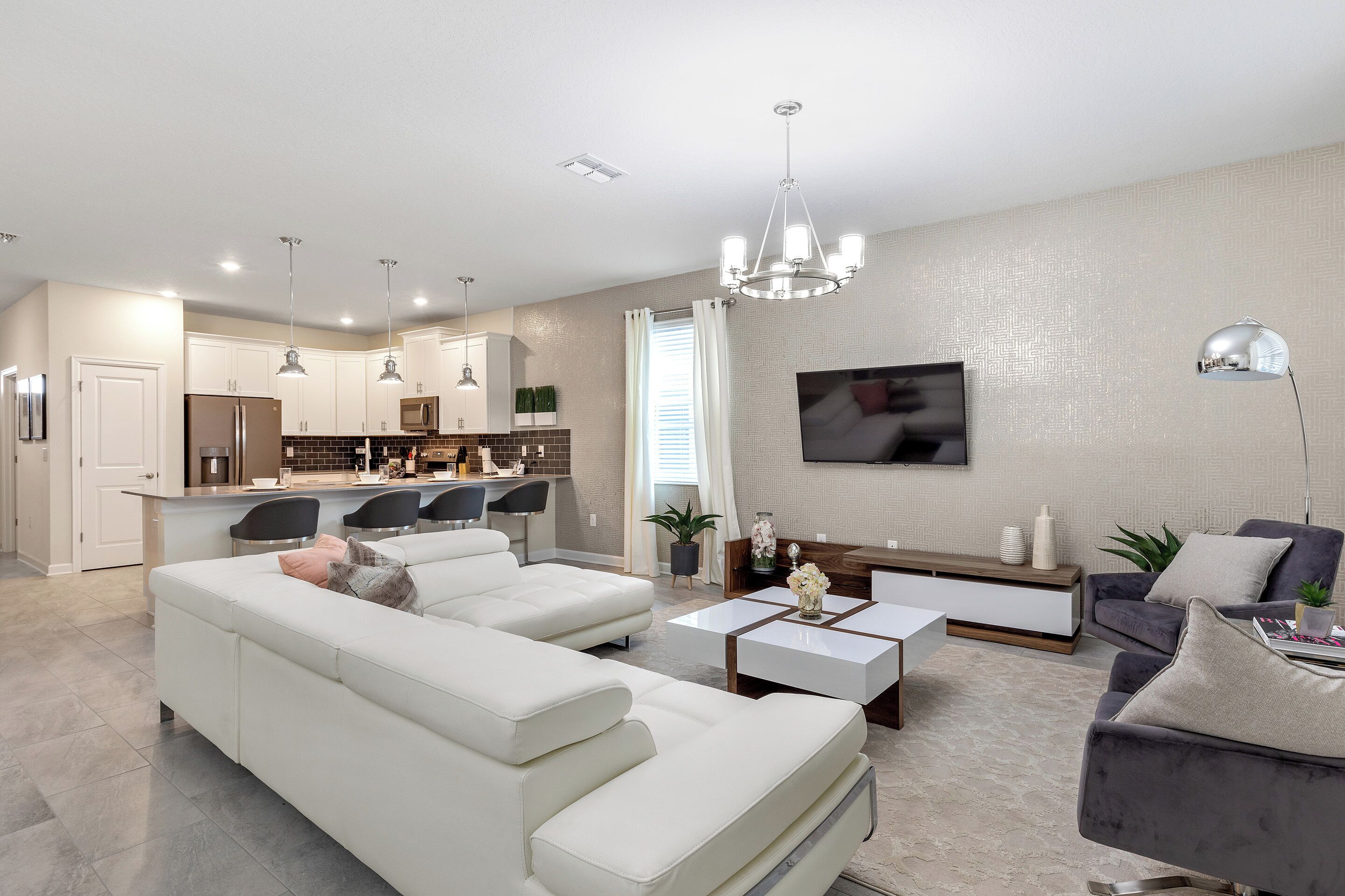 A modern living room with a white sofa, a coffee table, and a grey armchair. There is a kitchen in the background with white cabinets and a dining area.