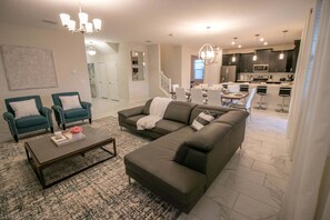 Uma sala de estar moderna com um sofá seccional, duas poltronas, uma mesa de centro e uma área de jantar, com uma cozinha ao fundo.