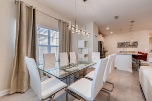 A modern dining area with a glass table and white chairs, a kitchen with a red bar seating, and a large window with blinds. - 4825 LL - Storey Lake - 5BD/4BA Private Pool (Kissimmee)