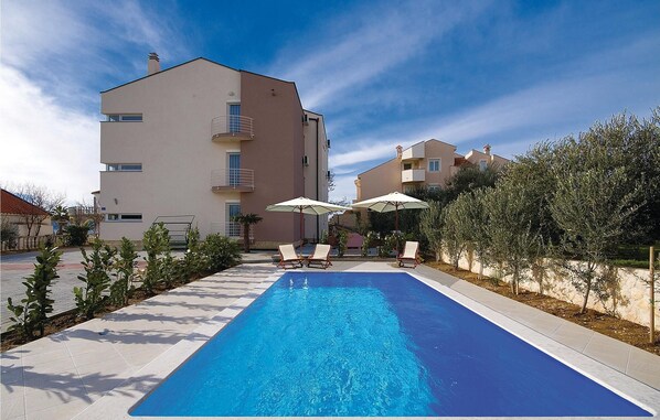 A swimming pool with lounge chairs and umbrellas, surrounded by a building with balconies and a clear sky.