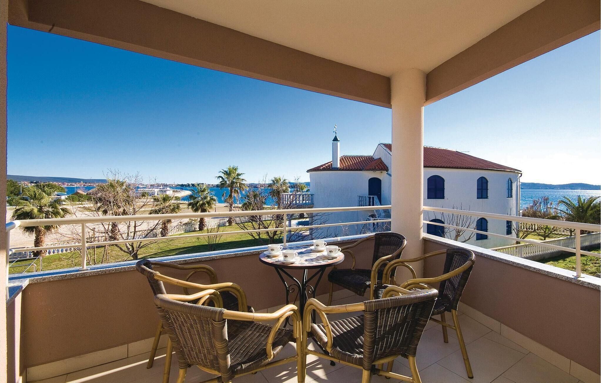 A balcony with wicker chairs and a table, overlooking a coastal view with buildings and palm trees.