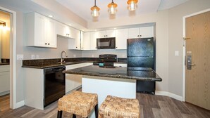 A modern kitchen with white cabinets, a granite countertop, a built-in microwave, a black refrigerator, and a central island with stools. - Renovated Penthouse Studio (Kihei)