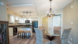 A modern kitchen with white cabinets, a central island, bar stools, and a glass dining table with wicker chairs. - Renovated Penthouse Studio (Kihei)