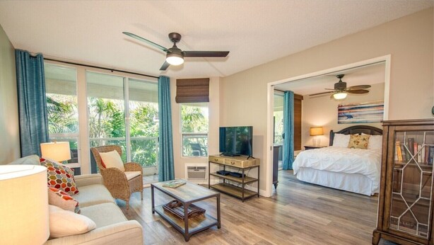 A spacious living room with a sofa, coffee table, and TV, leading to a bedroom with a bed and a ceiling fan. - Renovated Penthouse Studio (Kihei)
