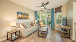 A living room with a sofa, coffee table, and TV stand. There is a ceiling fan and a window with curtains. - Renovated Penthouse Studio (Kihei)