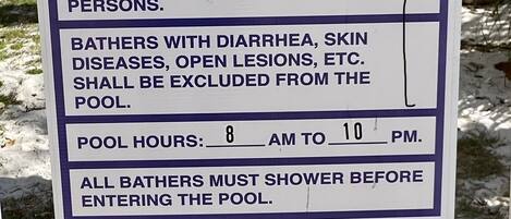 Letrero de normas de la piscina: aforo máximo de 25 personas, prohibido llevar vidrio o animales, exclusión de bañistas con ciertas condiciones de salud, horario de apertura de 8 de la mañana a 10 de la noche, y prohibido bucear.
