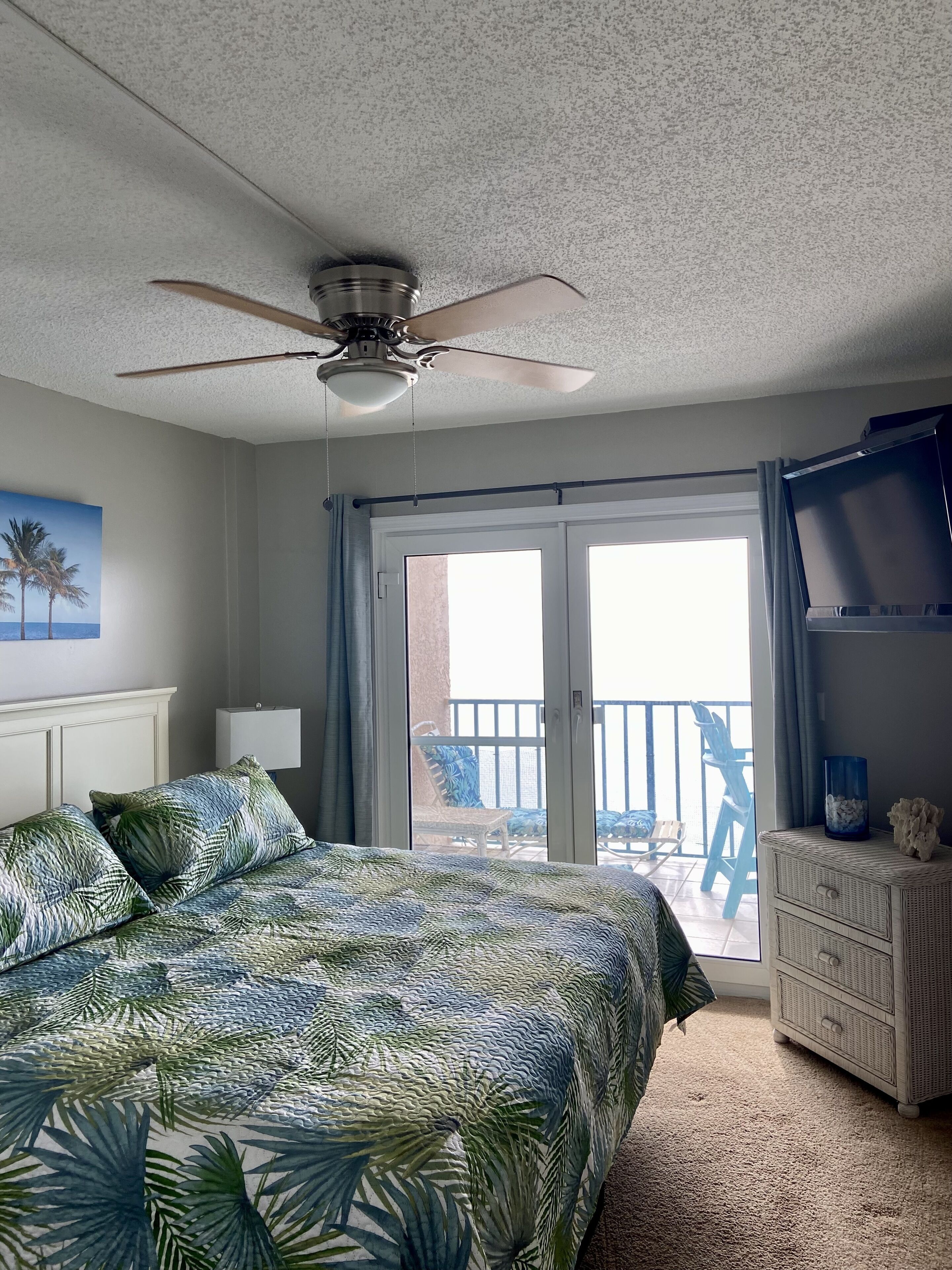 Une chambre avec un lit, un ventilateur de plafond, un balcon avec des chaises et une vue sur l’océan.