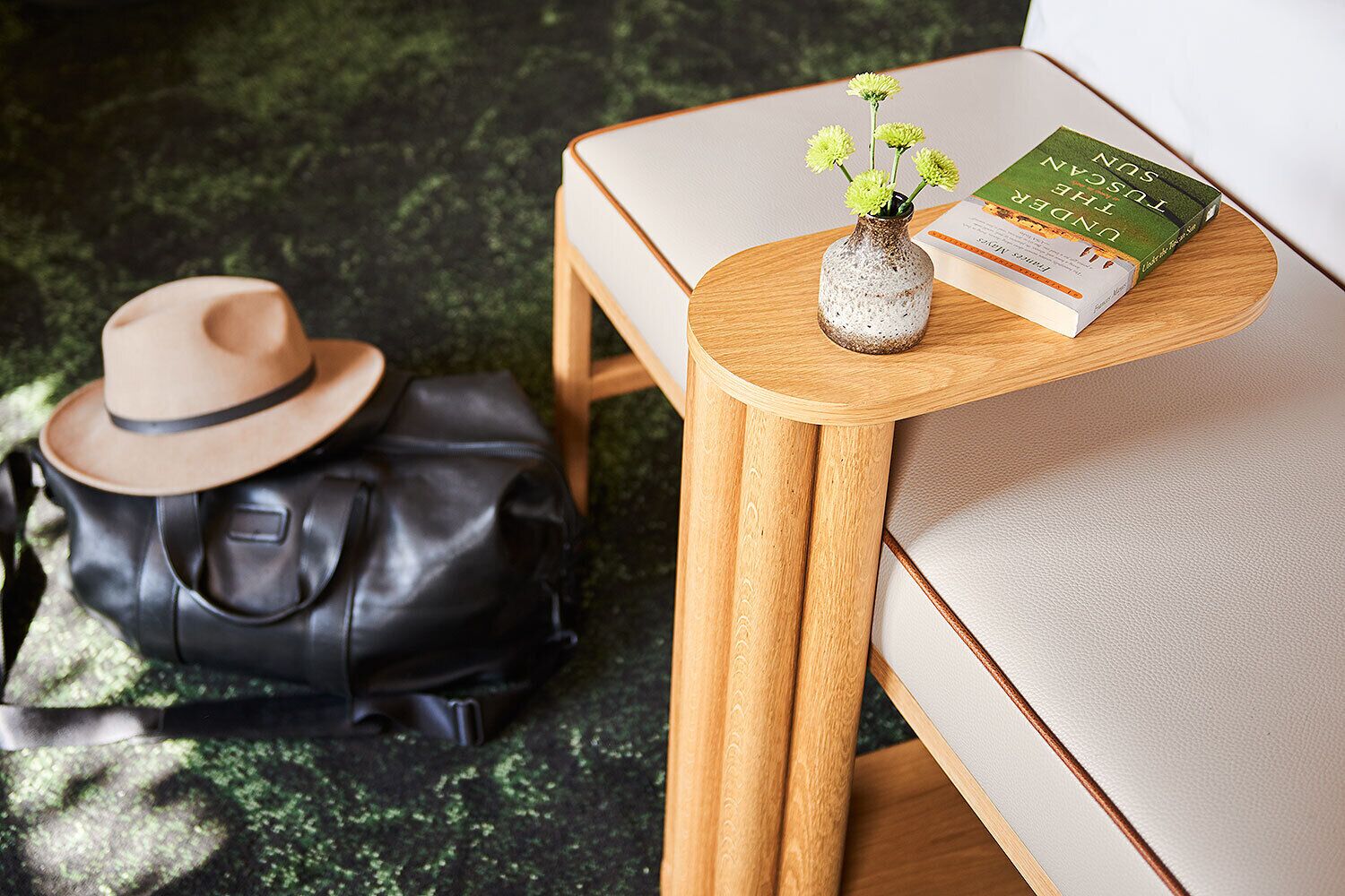 A wooden side table with a vase of flowers and a book on it, a leather bag, and a hat on the floor.