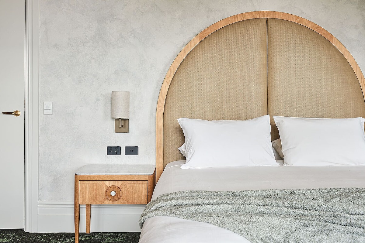 A bedroom with a large bed, a wooden nightstand with a drawer, a wall-mounted lamp, and a textured wall.