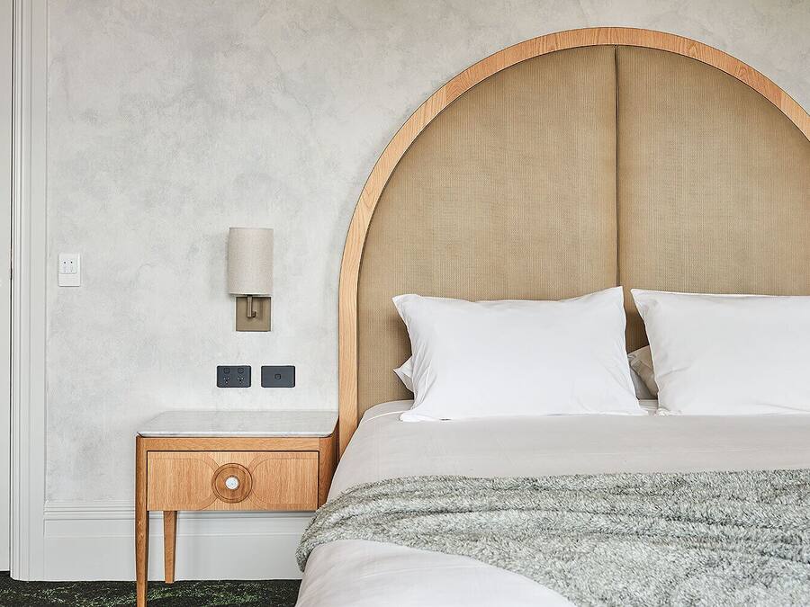 Un dormitorio con una cama grande, una mesita de noche de madera con cajón, una lámpara de pared y una pared con textura.