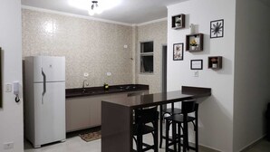 A compact kitchen with a refrigerator, stove, and sink. There are two bar stools and a small dining table. The walls are decorated with framed pictures and a flower arrangement. - Spacious, beautiful apartment - 3 blocks from the beach! (Guarujá)