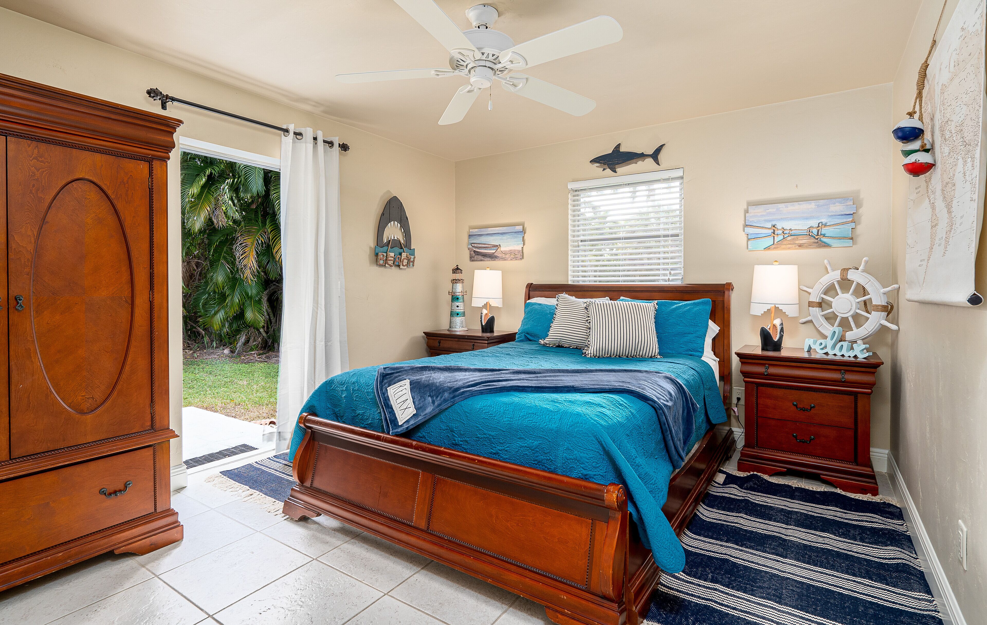 A bedroom with a wooden bed, a ceiling fan, a nightstand with a lamp, and a view of the outdoors.