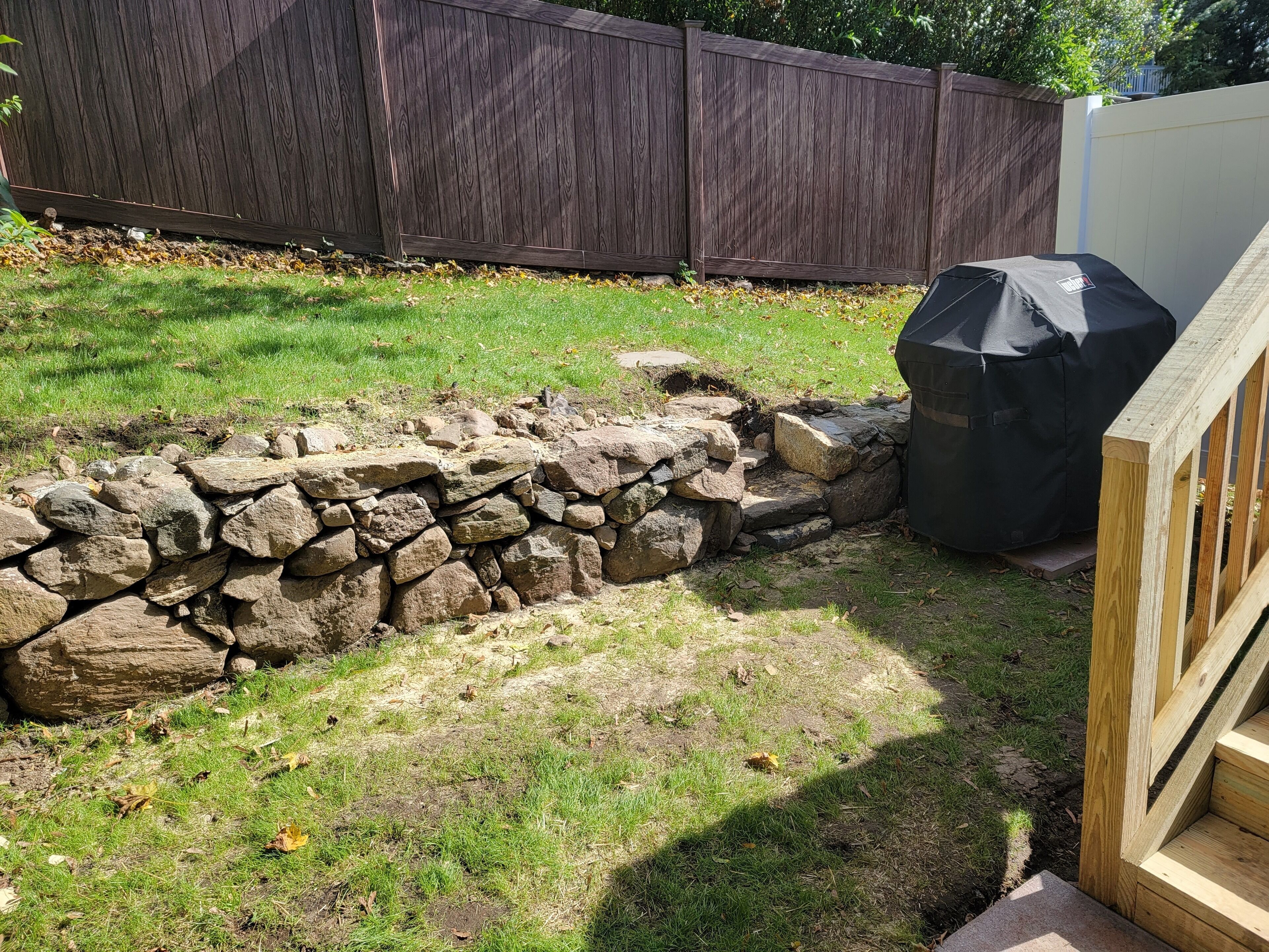 A backyard with a stone wall, a wooden deck, and a covered grill area.