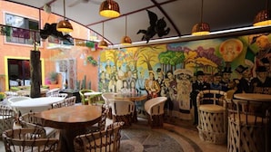 Ein Restaurant mit runden Tischen, Korbstühlen, Wandgemälde und hängenden Pendelleuchten.