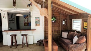 A rustic wooden interior with a bar area, wooden stools, a wicker sofa with cushions, and a wooden ceiling structure.