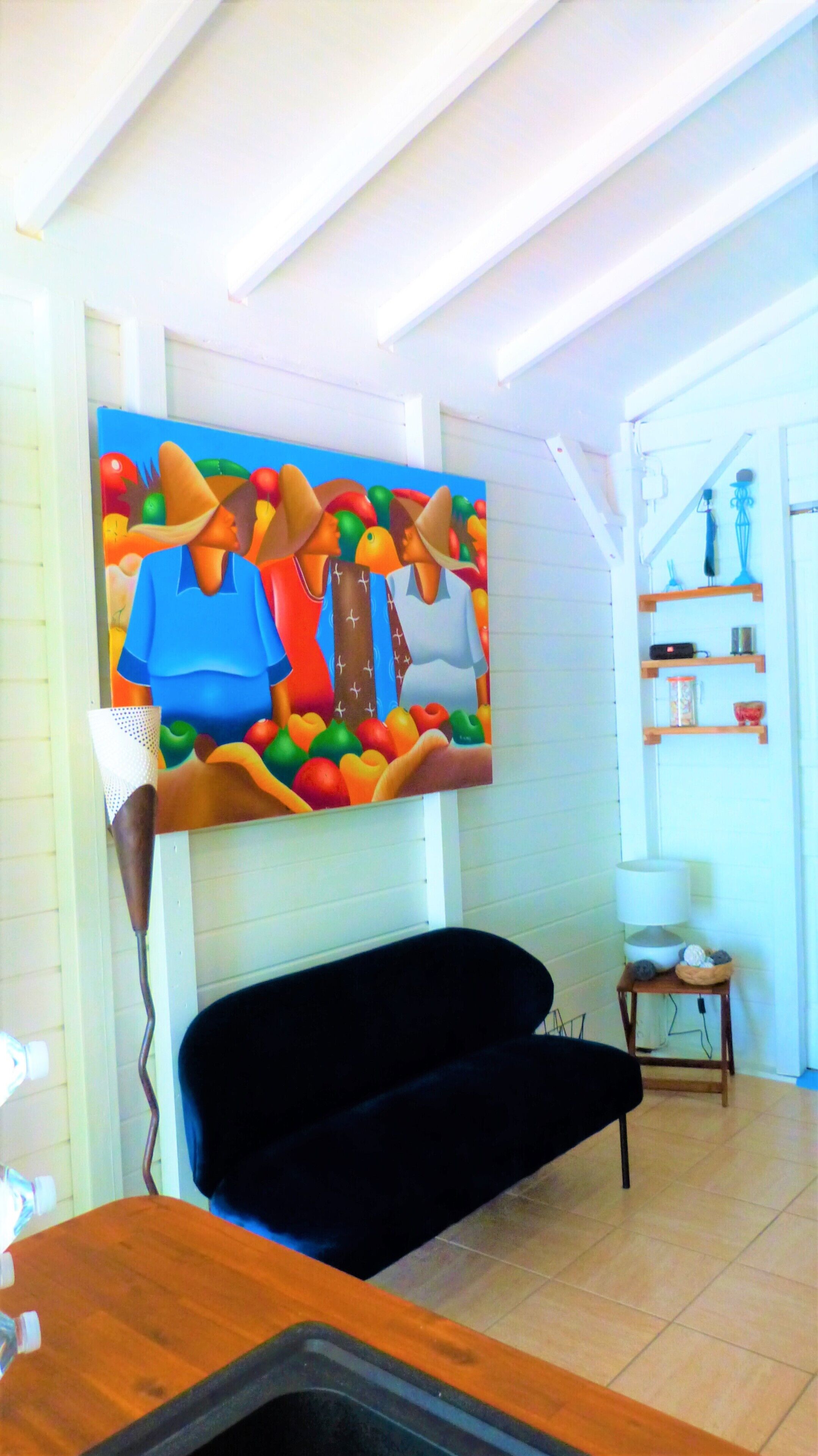 Une pièce avec un canapé noir, un tableau abstrait aux couleurs vives accroché au mur, une petite table en bois avec une lampe et une étagère ornée d’objets décoratifs.