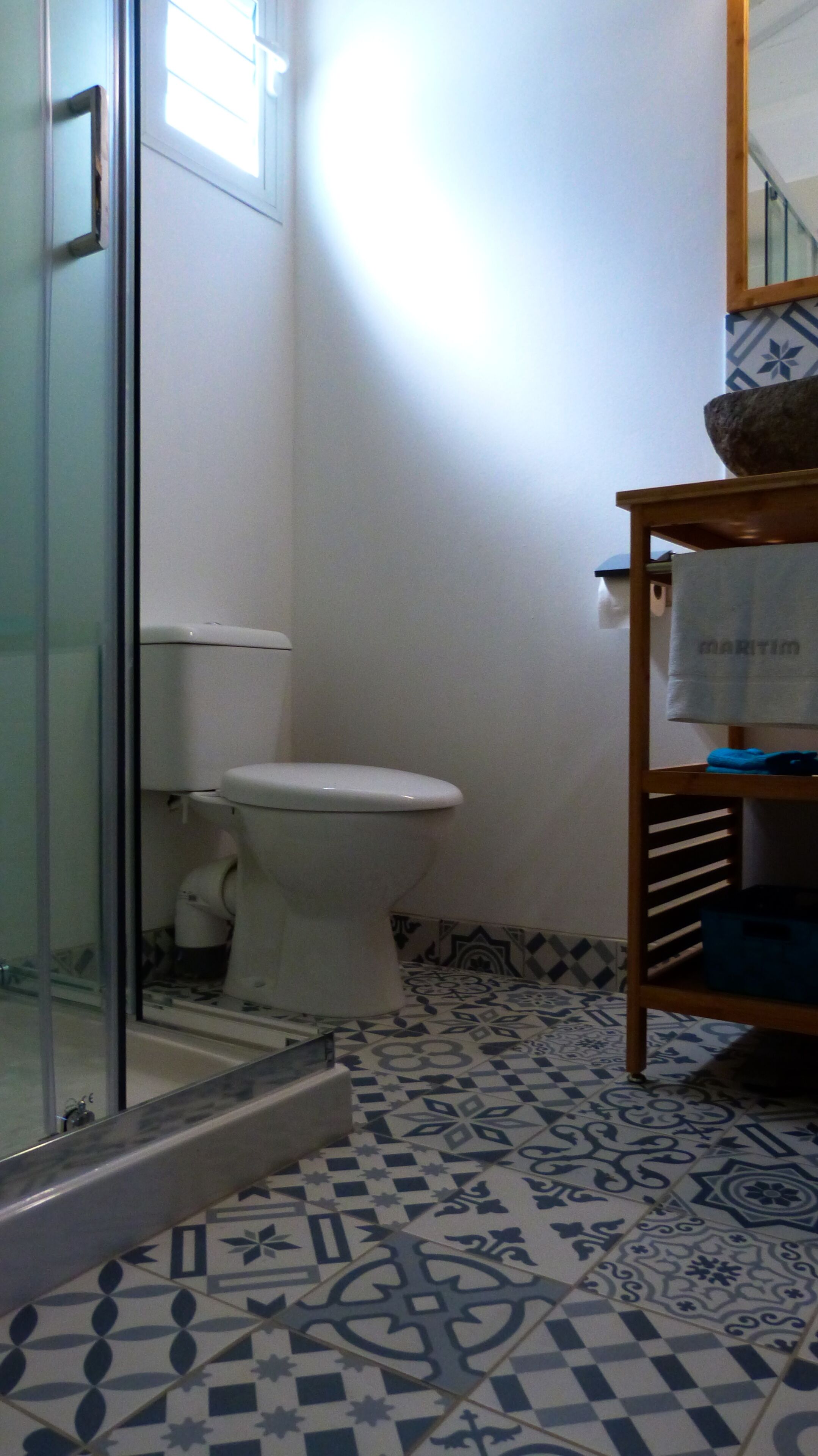 Un baño con inodoro, cabina de ducha de vidrio, suelo con baldosas estampadas y estante de madera con una toalla.