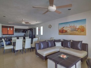 A modern living room with a sectional sofa, a dining area with chairs, a kitchen in the background, and ceiling fans.