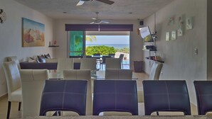 A modern living room with a large window overlooking a scenic view, a ceiling fan, and a dining area with chairs and a table. - Casa Nazul  -  Yucatan Home Rentals (Chicxulub Puerto)