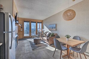 Una cocina moderna con horno empotrado y refrigerador, un comedor con mesa de madera y sillas grises, y un rincón de descanso acogedor con sofá y cojines.