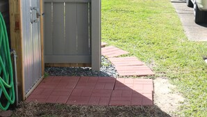 Un patio trasero cercado con manguera, un sendero de ladrillo y un coche estacionado.