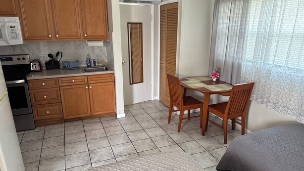 A compact kitchen with wooden cabinets, a microwave, and a toaster. There's a dining area with a table and chairs, and a bed with a grey blanket.