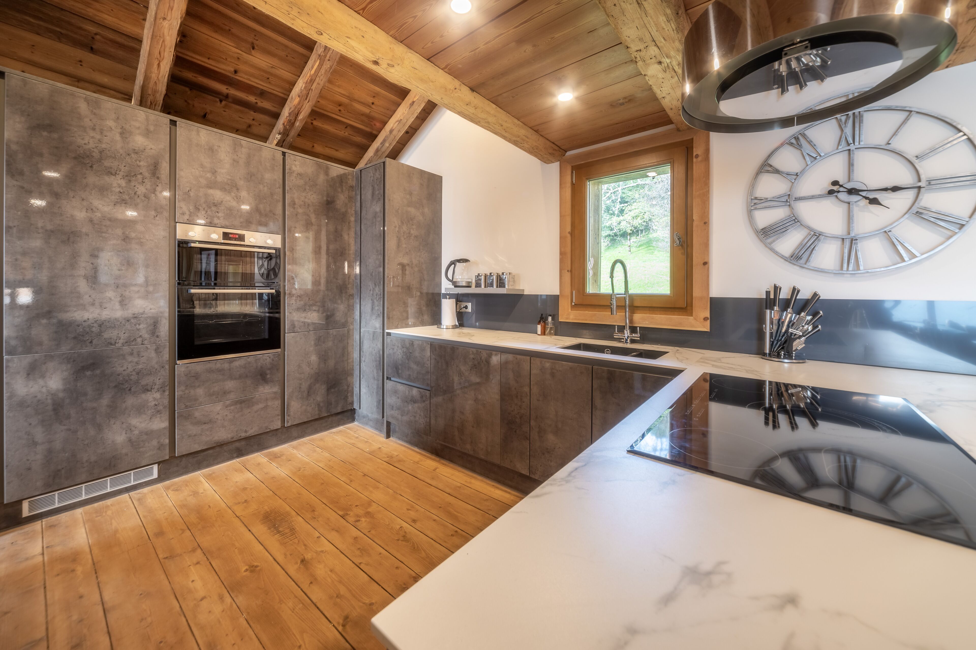 A modern kitchen with a built-in oven, a large clock, and a window providing natural light.