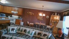 A cozy living room with a sofa featuring a bear and tree pattern, a dining area with a table and chairs, and a kitchen with wooden cabinets and a microwave.