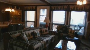 A cozy living room with a plaid sofa, a coffee table, and a fireplace. There are two windows with curtains, a lamp, and a bookshelf. - Cedar Village Condo A5, Beech Mountain, NC (Beech Mountain)