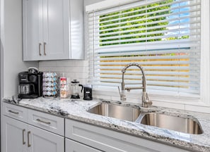 A kitchen with a double sink, a coffee maker, a grinder, and a window with blinds.