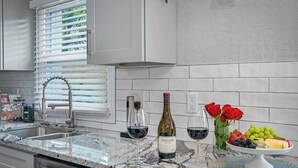 Een moderne keuken met een granieten aanrecht, een roestvrijstalen spoelbak en witte kasten. Op het aanrecht staan twee wijnflessen en glazen, samen met een schaal fruit met onder andere druiven en aardbeien.