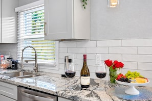 A modern kitchen with a granite countertop, stainless steel sink, and white cabinetry. There are two wine bottles, a glass of red wine, and a bowl of fruit on the counter.