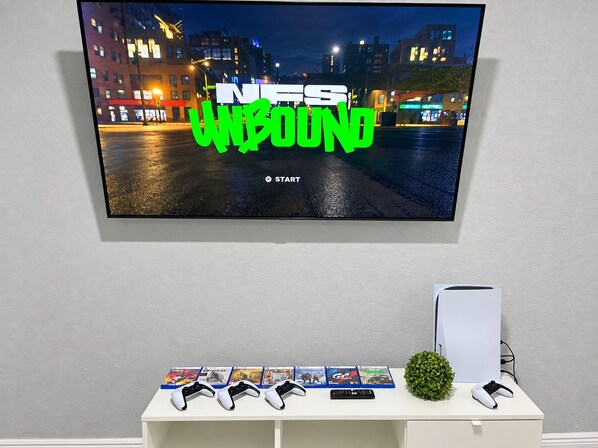 Un téléviseur fixé au mur affiche un jeu vidéo avec le texte « NFS Unbound » et un bouton « DÉMARRER ». Sous le téléviseur, on trouve une étagère blanche contenant des boîtiers de jeux vidéo, des manettes et une petite plante verte.