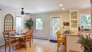 A dining area with wooden chairs and a table, a ceiling fan, and a kitchen with cabinets and a window. - Peaceful retreat plus Dog Friendly! (Wellfleet)