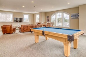 Une table de billard dans une pièce dont le sol est recouvert d’une moquette beige, avec un canapé en cuir marron et une table à manger entourée de chaises.