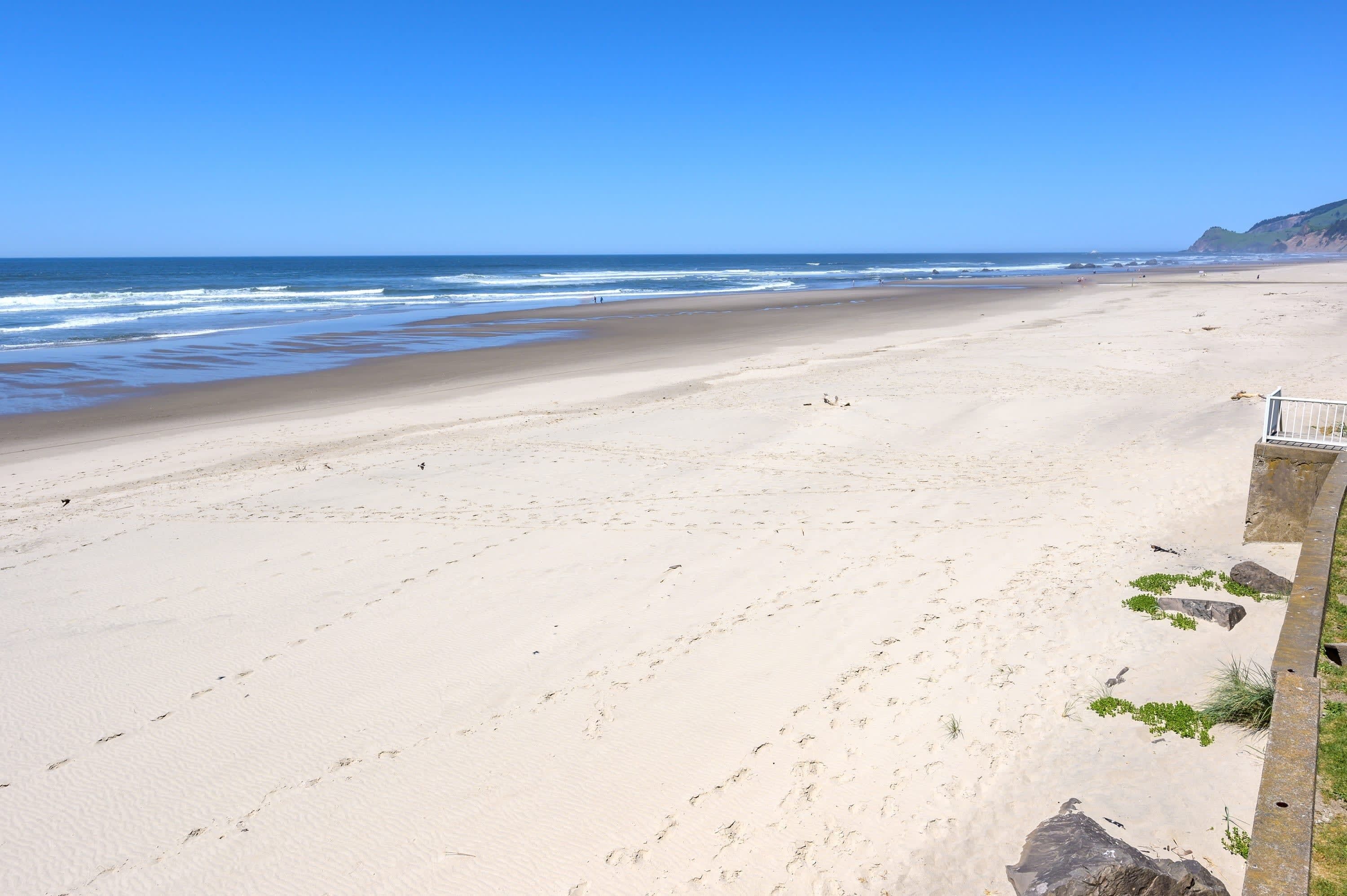 A wide sandy beach with gentle waves, a clear blue sky, and a rocky cliff in the distance.