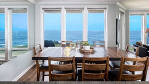 A dining room with a large table and chairs, large windows with ocean views, and a ceiling light fixture. - Coastal Breakers Condo, Ocean Patio, BBQ & Pool (Depoe Bay)