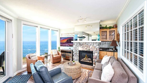 A spacious living room with a stone fireplace, a television, and a view of the ocean. - Coastal Breakers Condo, Ocean Patio, BBQ & Pool (Depoe Bay)