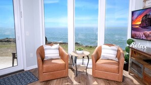 A room with a large window overlooking the ocean, two leather chairs, a small table with a plant, and a TV on a wooden stand. - Coastal Breakers Condo, Ocean Patio, BBQ & Pool (Depoe Bay)