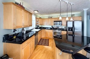 A modern kitchen with wooden cabinets, a black countertop, stainless steel appliances, and pendant lighting. - Captains Choice Condo, Panoramic Views & Pool (Depoe Bay)