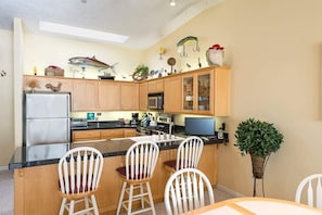 Een keuken met houten kastjes, een koelkast, een magnetron en een eethoek met witte stoelen.