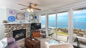 Ein Wohnzimmer mit Kamin aus Stein, Deckenventilator und großem Fenster mit Meerblick.