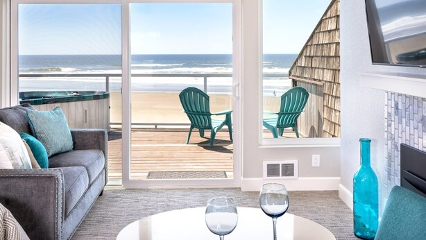 A room with a large sliding glass door leading to a balcony with a view of the ocean. There is a gray sofa with teal cushions, a round table with two glasses, and a blue vase.