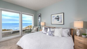 Une chambre avec un grand lit, deux tables de chevet, une chaise, une vue sur l’océan et un tableau encadré au mur.