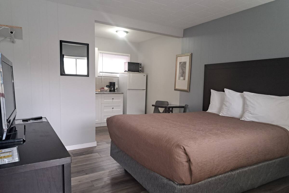 A bedroom with a bed, a desk, a chair, a small table, a refrigerator, a microwave, a coffee maker, and a window with blinds.
