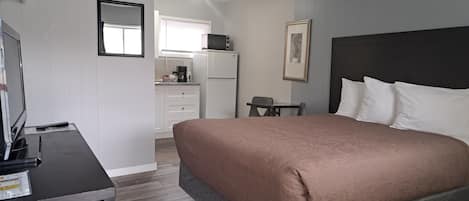 A bedroom with a bed, a desk, a chair, a small table, a refrigerator, a microwave, a coffee maker, and a window with blinds.