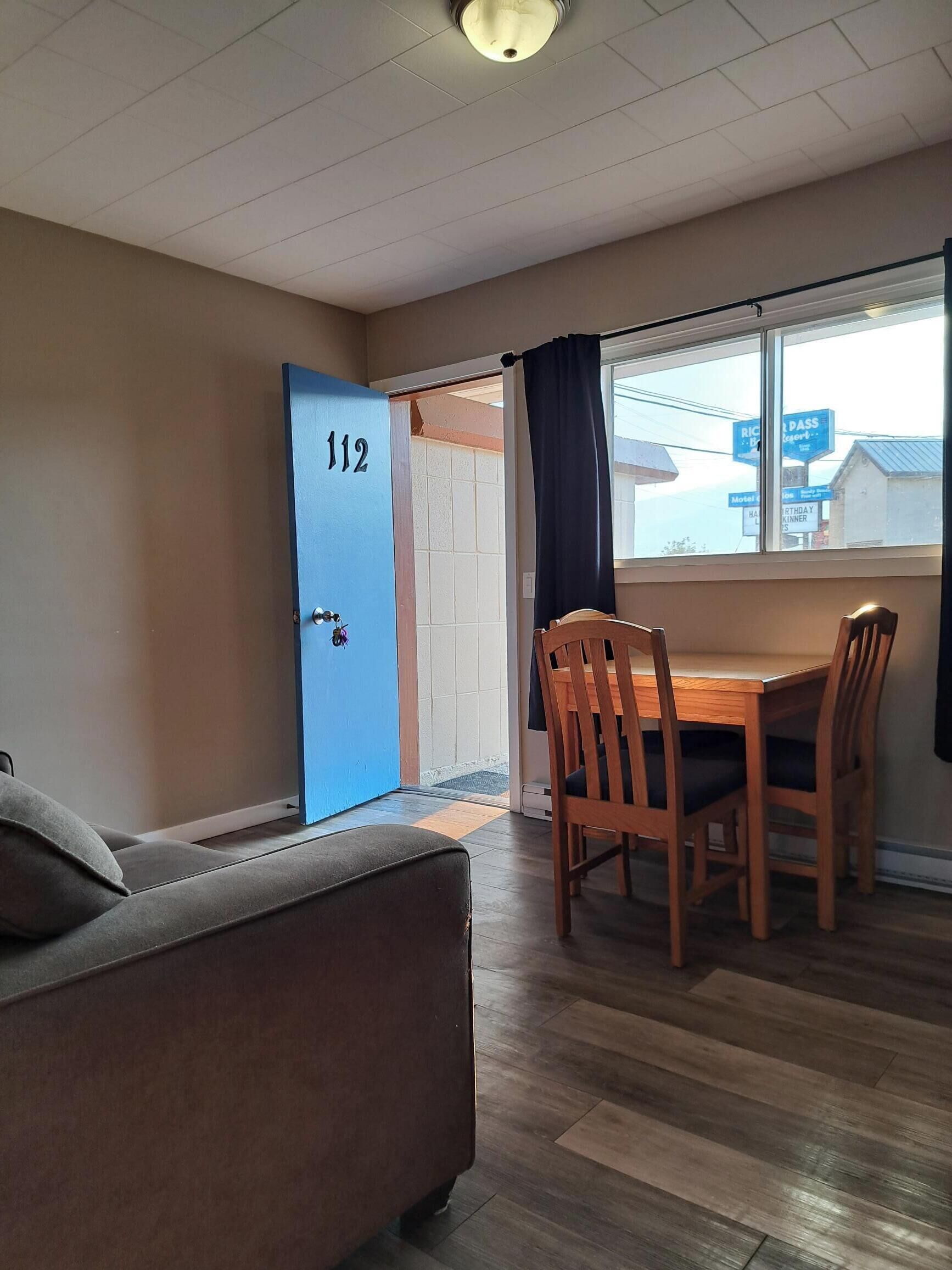 A room with a blue door marked 112, a dining table with chairs, a sofa, and a window with a view of buildings.