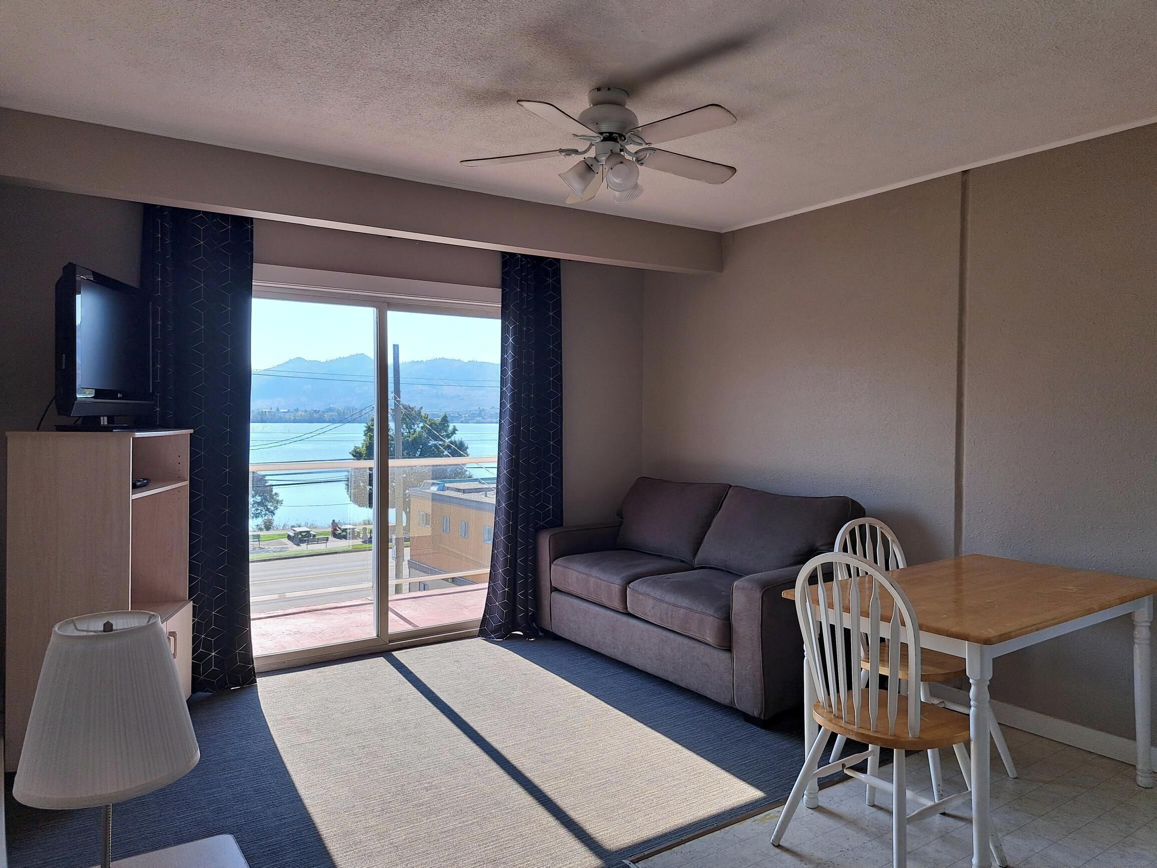 A room with a sofa, a table, a chair, a ceiling fan, a TV on a cabinet, and a view of a body of water and mountains.